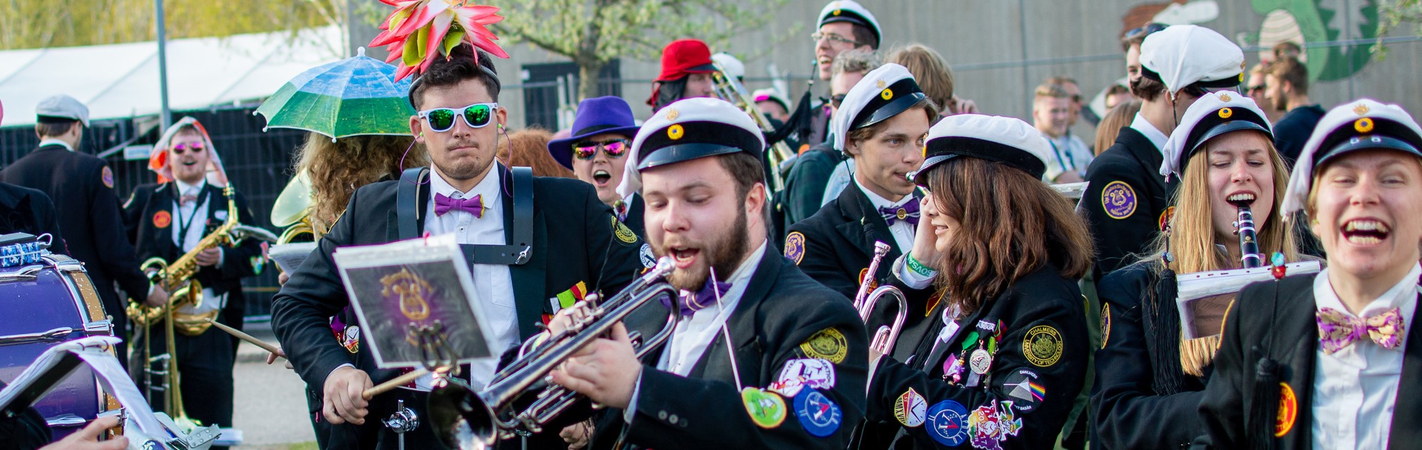 Studentorkester i parad med musiker i svarta kavajer, vita mössor och färgglada märken som spelar instrument och marscherar festligt